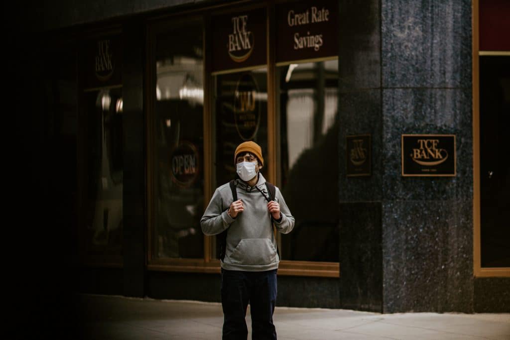 A person wearing a beanie, glasses, a face mask, and a hoodie stands on a sidewalk with a backpack. Behind them is a dark building with large windows and signs that read "ICE BANK" and "Great Rate Savings," while their phone's Exposure Notifications feature ensures timely health alerts.