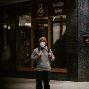 A person wearing a beanie, glasses, a face mask, and a hoodie stands on a sidewalk with a backpack. Behind them is a dark building with large windows and signs that read "ICE BANK" and "Great Rate Savings," while their phone's Exposure Notifications feature ensures timely health alerts.