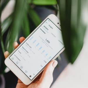 A hand holding a smartphone displaying Instagram app analytics data with bar graphs. The background is slightly blurred with green leaves, creating a natural setting, perfect for attracting new followers.