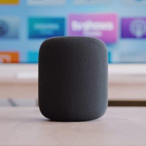tvOS 18: A black, cylindrical Apple HomePod Mini sits on a wooden surface with a television and its colorful app icons blurred in the background.