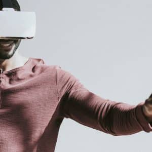 A person wearing a maroon shirt is using a virtual reality headset, possibly benefiting from innovative technology since Apple acquired NextVR. They are extending their arms forward as if interacting with a virtual environment, with a cheerful expression on their face. The background is plain and well-lit.