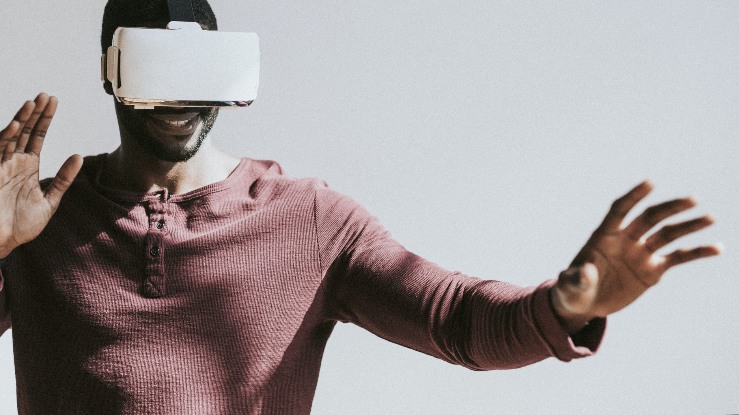 A person wearing a maroon shirt is using a virtual reality headset, possibly benefiting from innovative technology since Apple acquired NextVR. They are extending their arms forward as if interacting with a virtual environment, with a cheerful expression on their face. The background is plain and well-lit.