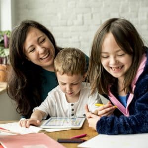 A woman and two children are sitting at a table, engaged in different activities. The boy is focusing on a tablet while the girl, holding a pencil, is smiling. The woman is also smiling and leaning towards the children. The room, suited for homeschooling during COVID-19, has a bright ambiance with a brick wall in the background.