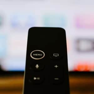 Close-up of a black remote control pointed towards a blurred television screen displaying colorful, indistinct icons. The remote features several buttons, including one labeled "MENU," a circular button with a rectangle icon, and volume control. Perfectly compatible with your new 4K Apple TV.