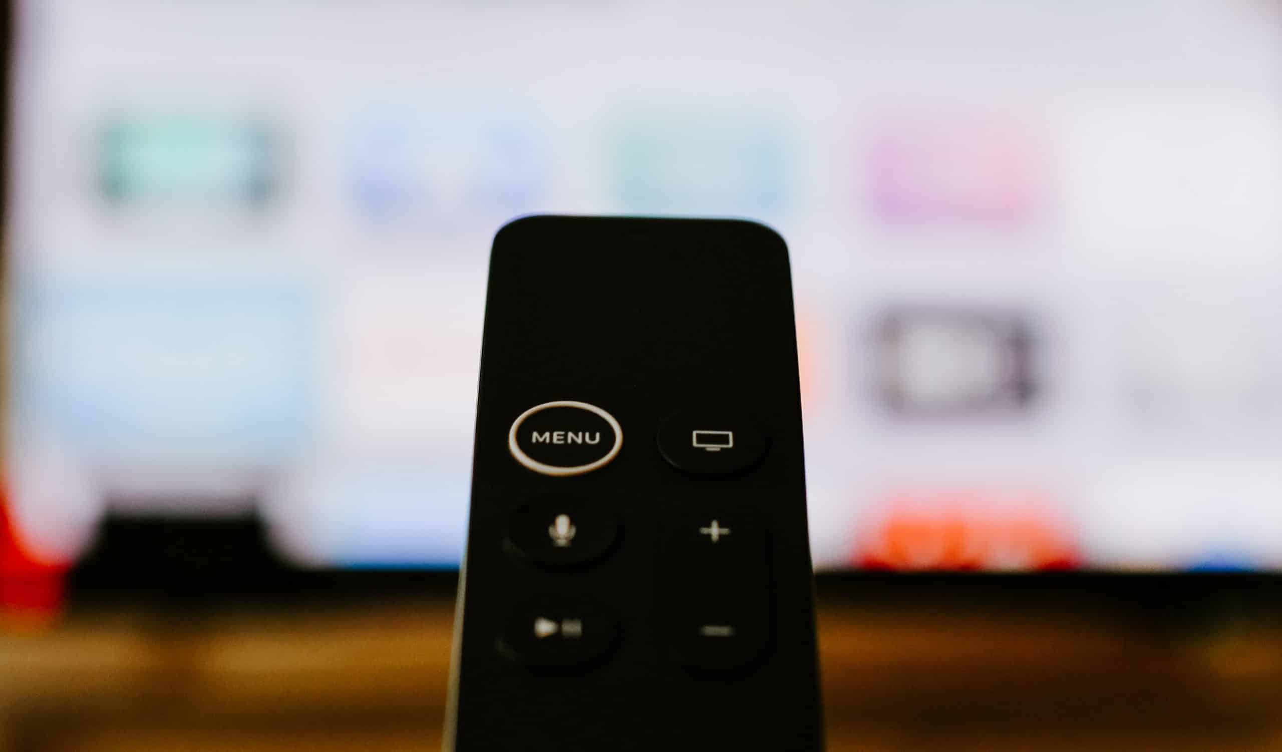 Close-up of a black remote control pointed towards a blurred television screen displaying colorful, indistinct icons. The remote features several buttons, including one labeled "MENU," a circular button with a rectangle icon, and volume control. Perfectly compatible with your new 4K Apple TV.