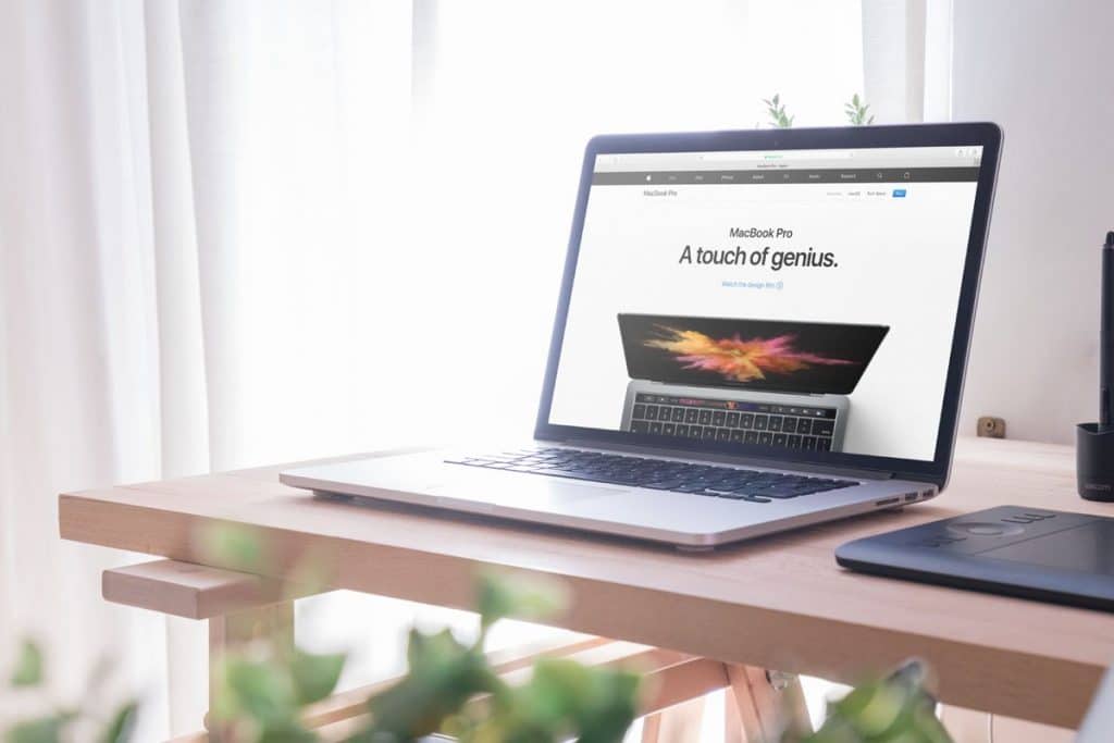 A MacBook Pro is shown on a wooden desk near a window with sheer white curtains. The laptop screen displays an advertisement that reads "MacBook Pro A touch of genius." A green plant and a black mouse pad are also visible, creating an ideal workspace for programmers.