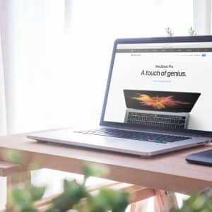 A MacBook Pro is shown on a wooden desk near a window with sheer white curtains. The laptop screen displays an advertisement that reads "MacBook Pro A touch of genius." A green plant and a black mouse pad are also visible, creating an ideal workspace for programmers.