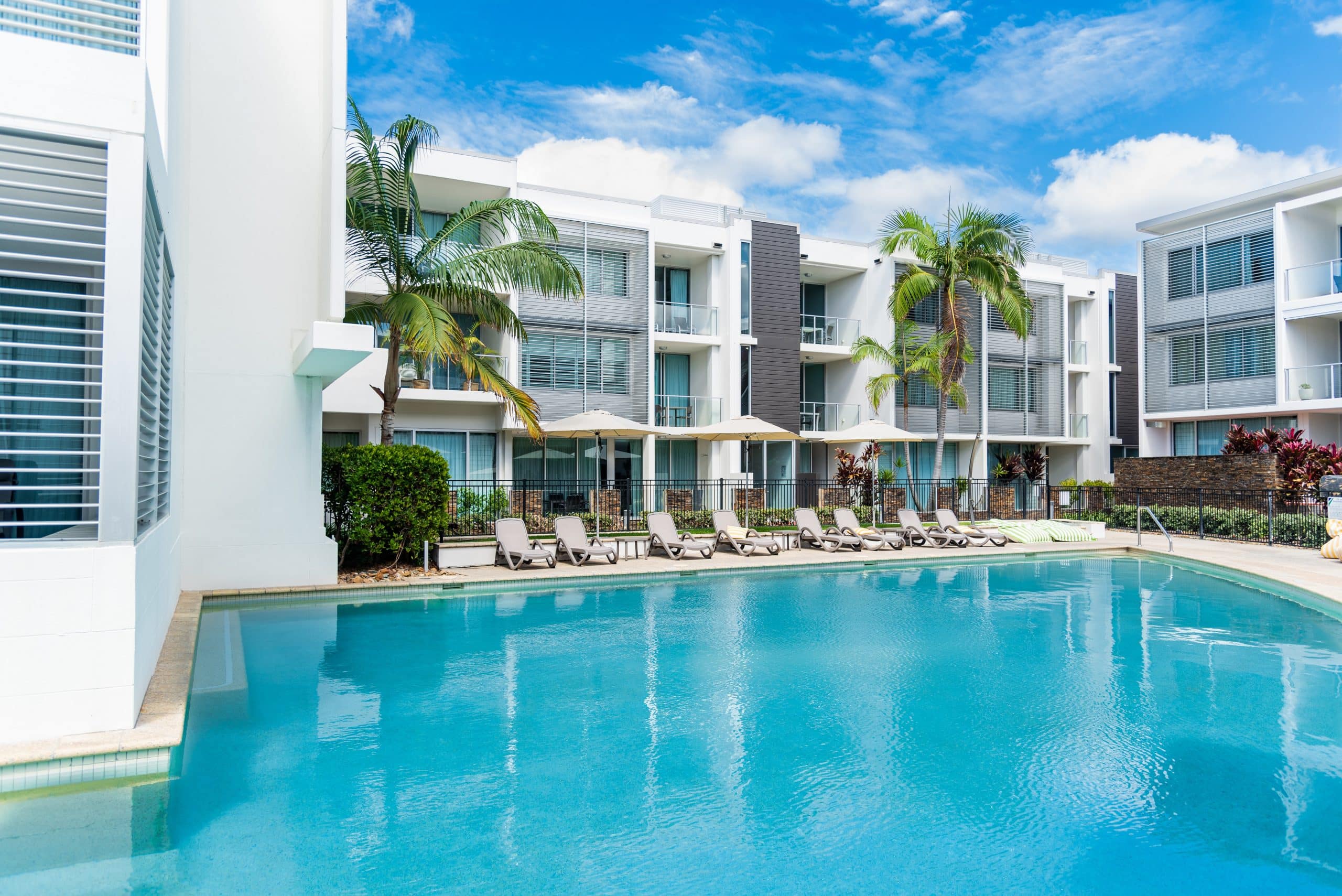 A modern outdoor swimming pool with clear blue water, surrounded by lounge chairs and umbrellas. The pool area at the new Apple Hotel in Austin, Texas is lined with white multi-story buildings featuring glass balconies and palm trees, creating a tropical and contemporary atmosphere under a bright blue sky.