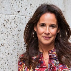 A person with long, dark hair and hoop earrings is standing against a textured wall, smiling slightly. They are wearing a colorful, patterned shirt with a high collar. The lighting is natural, and their expression appears calm and confident, like someone ready to introduce new content on Apple TV+.