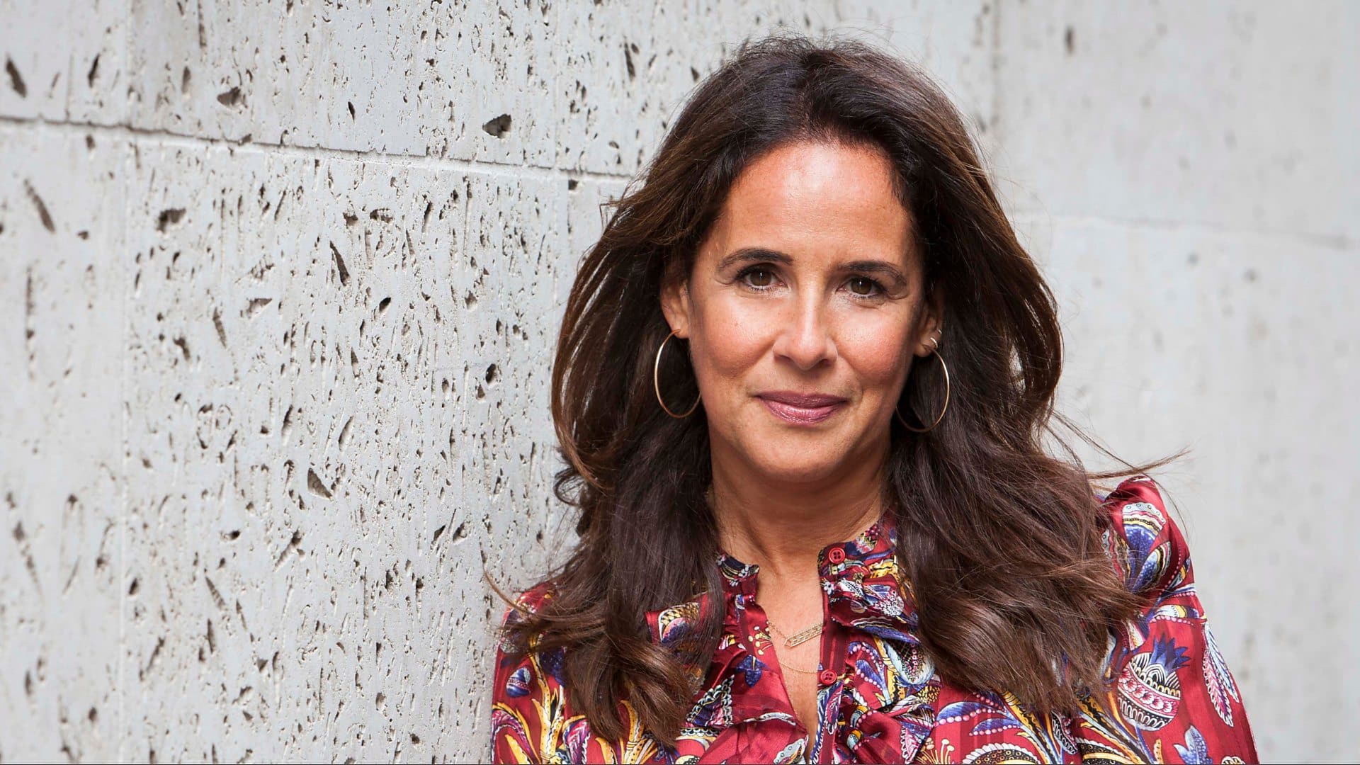 A person with long, dark hair and hoop earrings is standing against a textured wall, smiling slightly. They are wearing a colorful, patterned shirt with a high collar. The lighting is natural, and their expression appears calm and confident, like someone ready to introduce new content on Apple TV+.