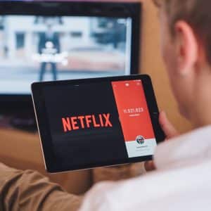 A person holds a tablet displaying the Netflix logo and a subscriber count while sitting on a couch. A TV in the background shows a blurred image. The person appears engaged with the screen on their tablet, possibly considering canceling inactive subscriptions to keep their account streamlined.