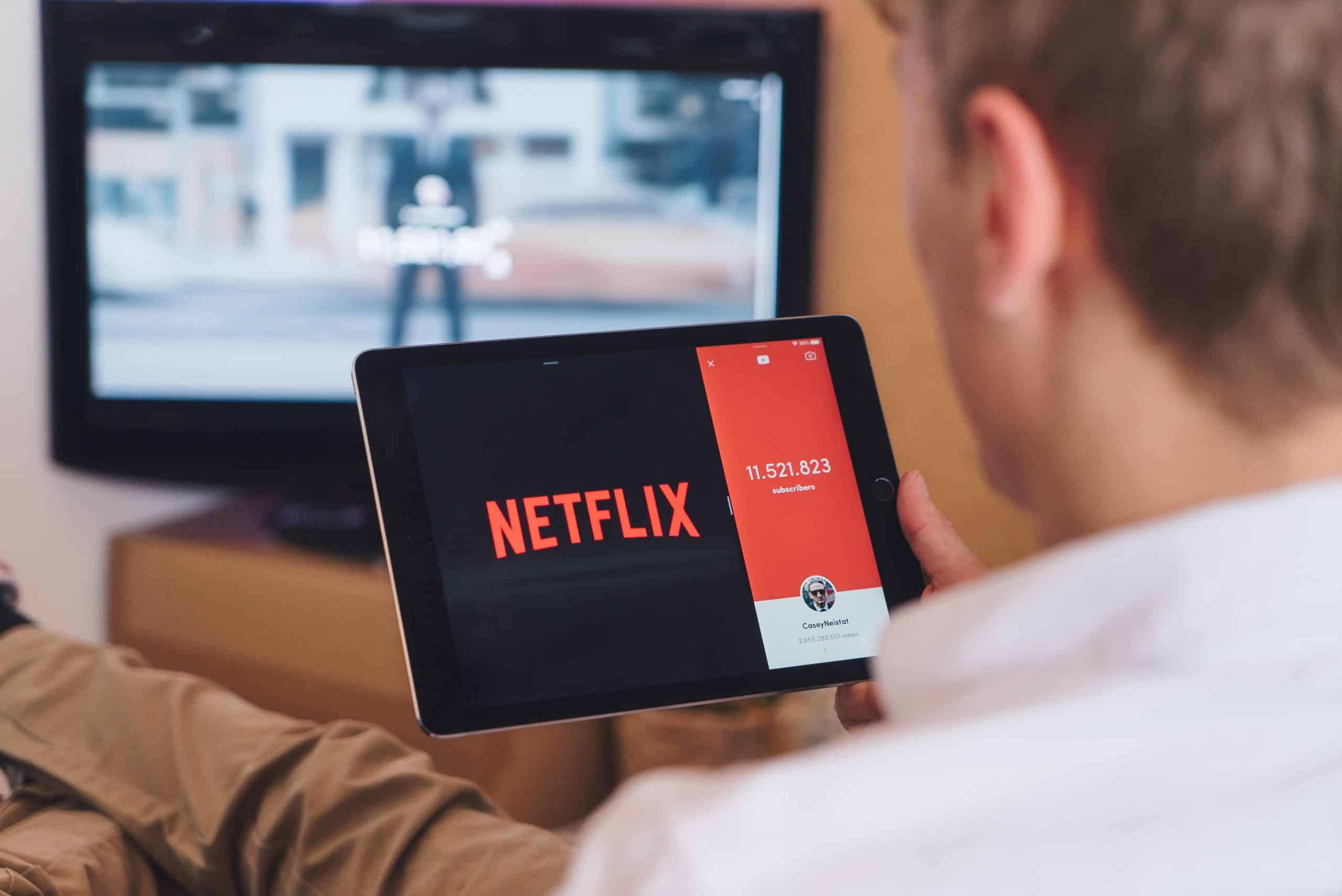 A person holds a tablet displaying the Netflix logo and a subscriber count while sitting on a couch. A TV in the background shows a blurred image. The person appears engaged with the screen on their tablet, possibly considering canceling inactive subscriptions to keep their account streamlined.