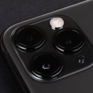 Close-up of the rear camera system on a modern smartphone. The image showcases three lenses and a flash, all housed in a sleek, dark-colored, square module on the phone's back surface. The smartphone, compatible with iOS 14, lies on a dark, textured background.