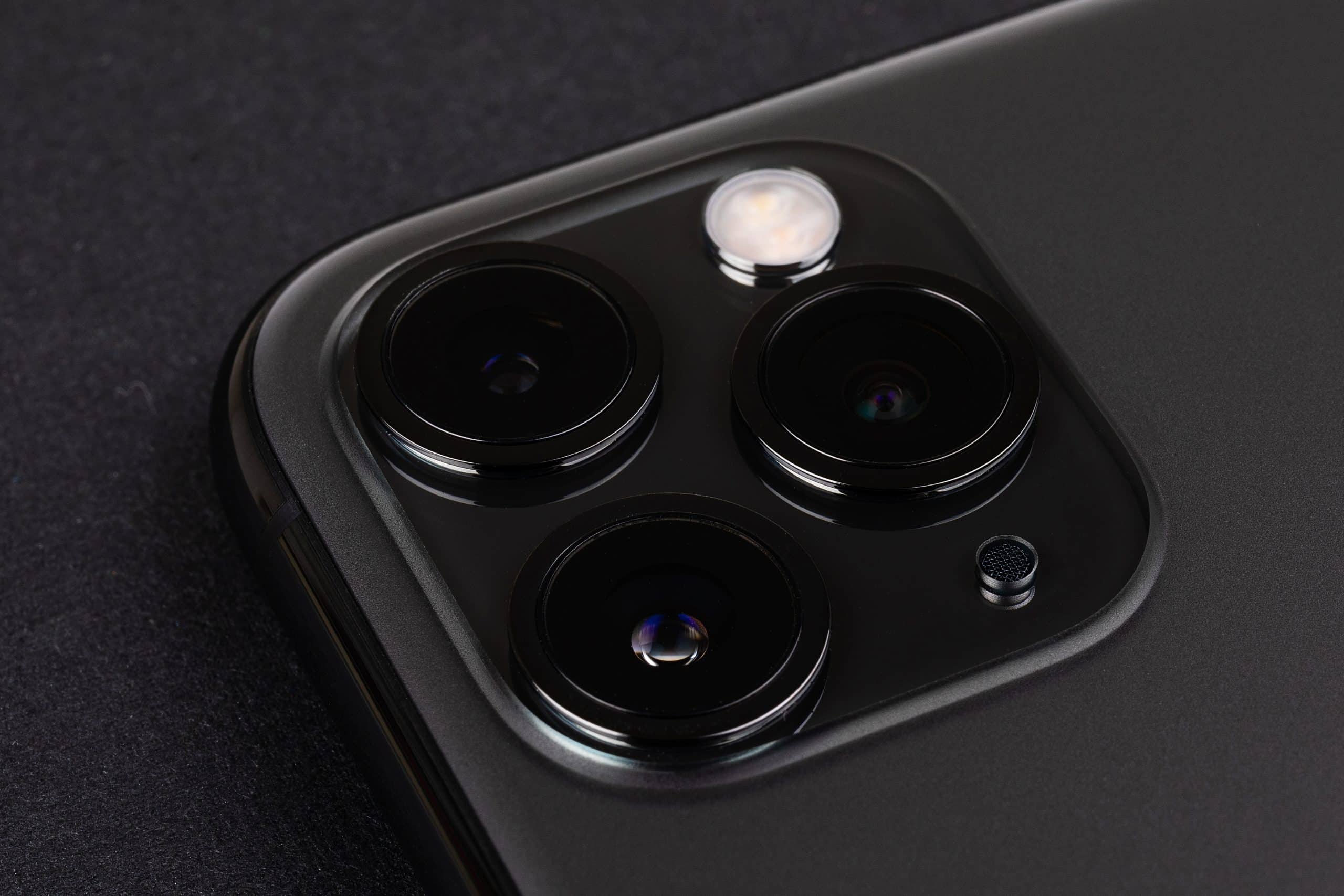 Close-up of the rear camera system on a modern smartphone. The image showcases three lenses and a flash, all housed in a sleek, dark-colored, square module on the phone's back surface. The smartphone, compatible with iOS 14, lies on a dark, textured background.