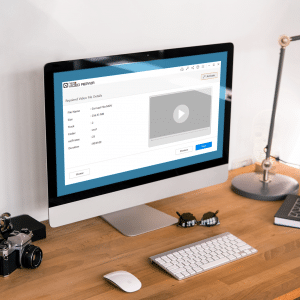 A desktop computer on a wooden desk displays a video editing software interface. Surrounding the computer are a camera, mouse, keyboard, pen, notepad, and various desk accessories. A lamp and some plants are also placed on the desk.