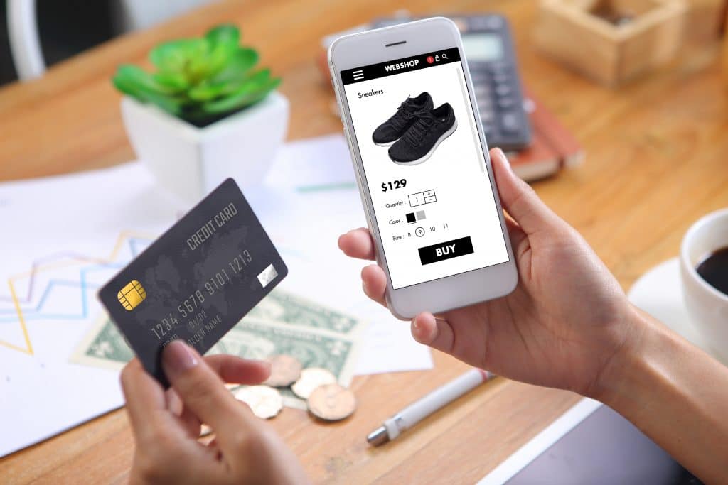 A person is holding a smartphone displaying an online shopping page for sneakers priced at $129. They hold a credit card in the other hand, ready to make the purchase. In the background, there's a potted plant, graph paper, coins, dollar bills, and a white cup—reflecting the booming sales contributing to $500 billion on platforms like the App Store.