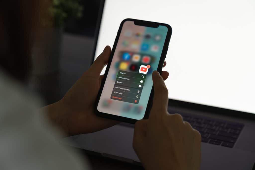 A person holds an iPhone displaying a contextual menu for the YouTube app, offering options like “Search,” “Subscriptions,” and “Create.” The background shows a blurred laptop screen. The person is using their other hand to interact with the phone, possibly browsing deals from the 6/18 shopping festival.