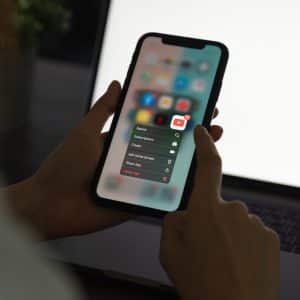 A person holds an iPhone displaying a contextual menu for the YouTube app, offering options like “Search,” “Subscriptions,” and “Create.” The background shows a blurred laptop screen. The person is using their other hand to interact with the phone, possibly browsing deals from the 6/18 shopping festival.