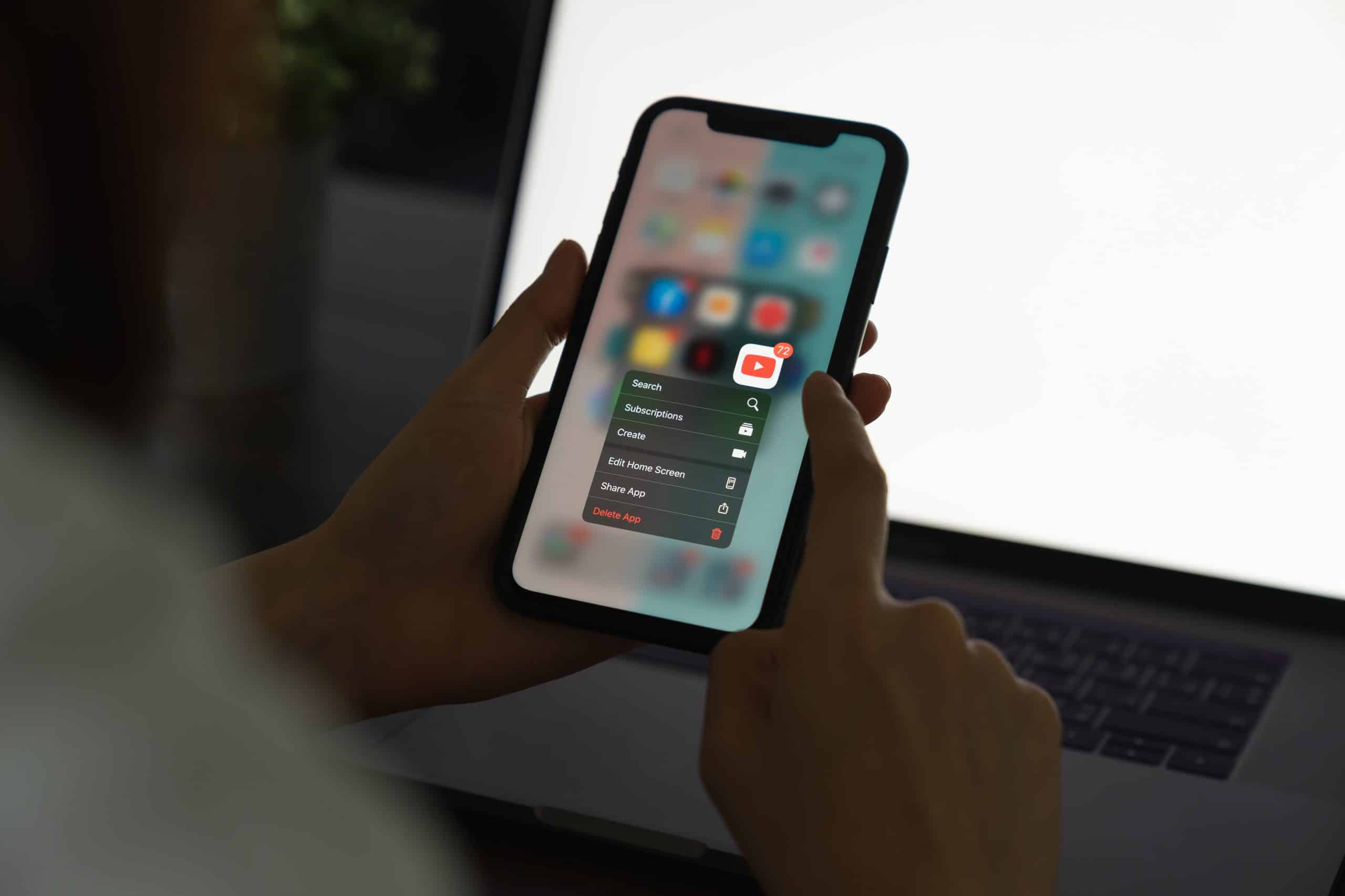 A person holds an iPhone displaying a contextual menu for the YouTube app, offering options like “Search,” “Subscriptions,” and “Create.” The background shows a blurred laptop screen. The person is using their other hand to interact with the phone, possibly browsing deals from the 6/18 shopping festival.