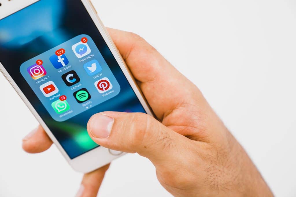 A person holding a smartphone displaying a screen with several social media and messaging apps including Instagram, WhatsApp, Telegram, Facebook Messenger, as well as other apps like Spotify, Pinterest, Camera, and a black C icon. The phone appears to have Touch ID for seamless security.