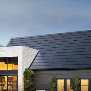 A modern house with new solar panels on its sleek roof, a mix of dark and light exterior walls, and large windows that give a glimpse of the warmly lit interior. Surrounding the house are green trees and shrubs under a clear sky.
