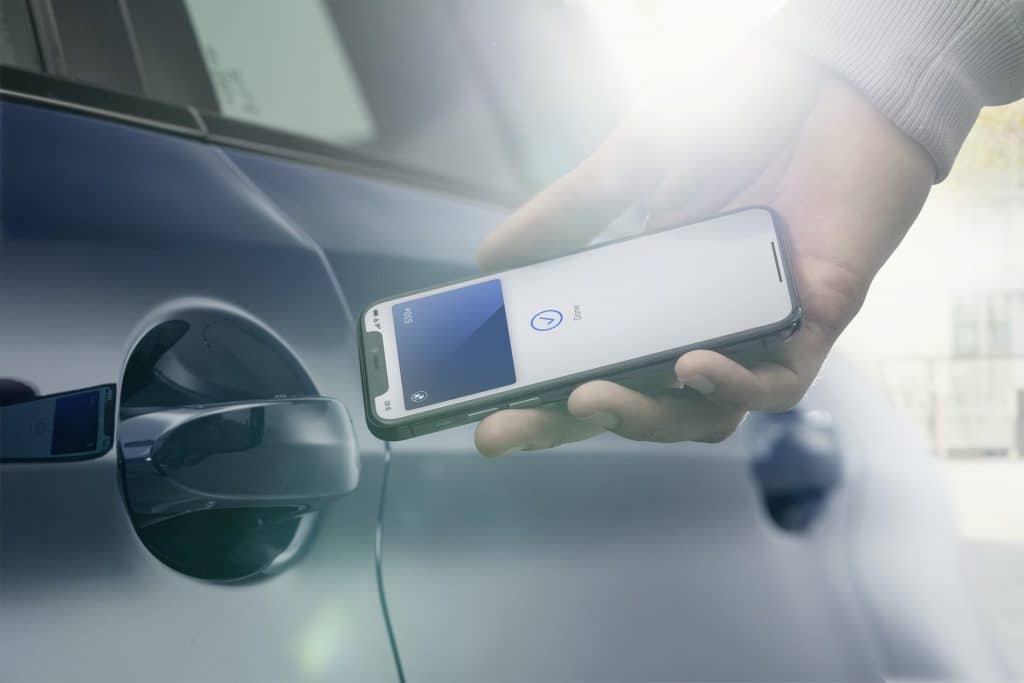 A person holds an iPhone near the door handle of a blue car. The phone screen displays the Apple CarKey app, suggesting the phone is being used to unlock the vehicle. Bright sunlight shines in the background.