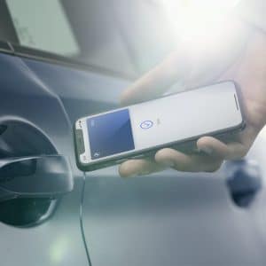 A person holds an iPhone near the door handle of a blue car. The phone screen displays the Apple CarKey app, suggesting the phone is being used to unlock the vehicle. Bright sunlight shines in the background.
