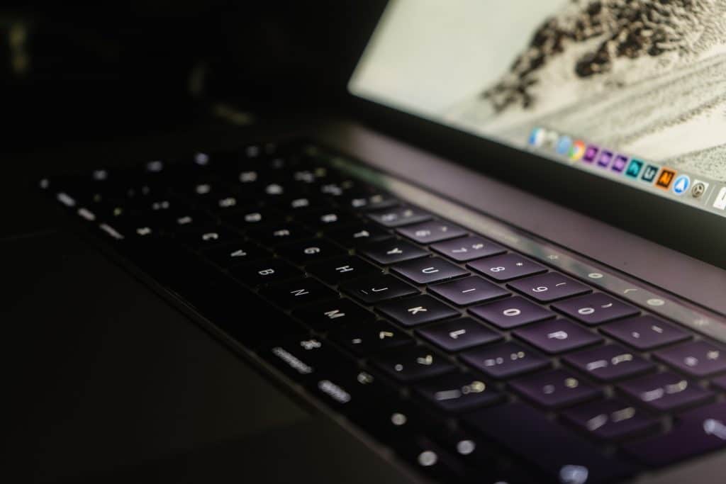 Close-up of an open Apple laptop with a backlit keyboard. The screen is partially visible, displaying various application icons and a WWDC announcement. The dimly lit environment highlights the glow from the keys and screen, creating a focused and slightly moody atmosphere.