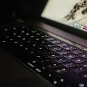 Close-up of an open Apple laptop with a backlit keyboard. The screen is partially visible, displaying various application icons and a WWDC announcement. The dimly lit environment highlights the glow from the keys and screen, creating a focused and slightly moody atmosphere.