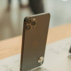 A modern Apple smartphone with a triple-camera setup is displayed on a wooden counter. The phone is standing upright, supported by a security stand. The background is slightly blurred, indicating an indoor environment, likely US stores.