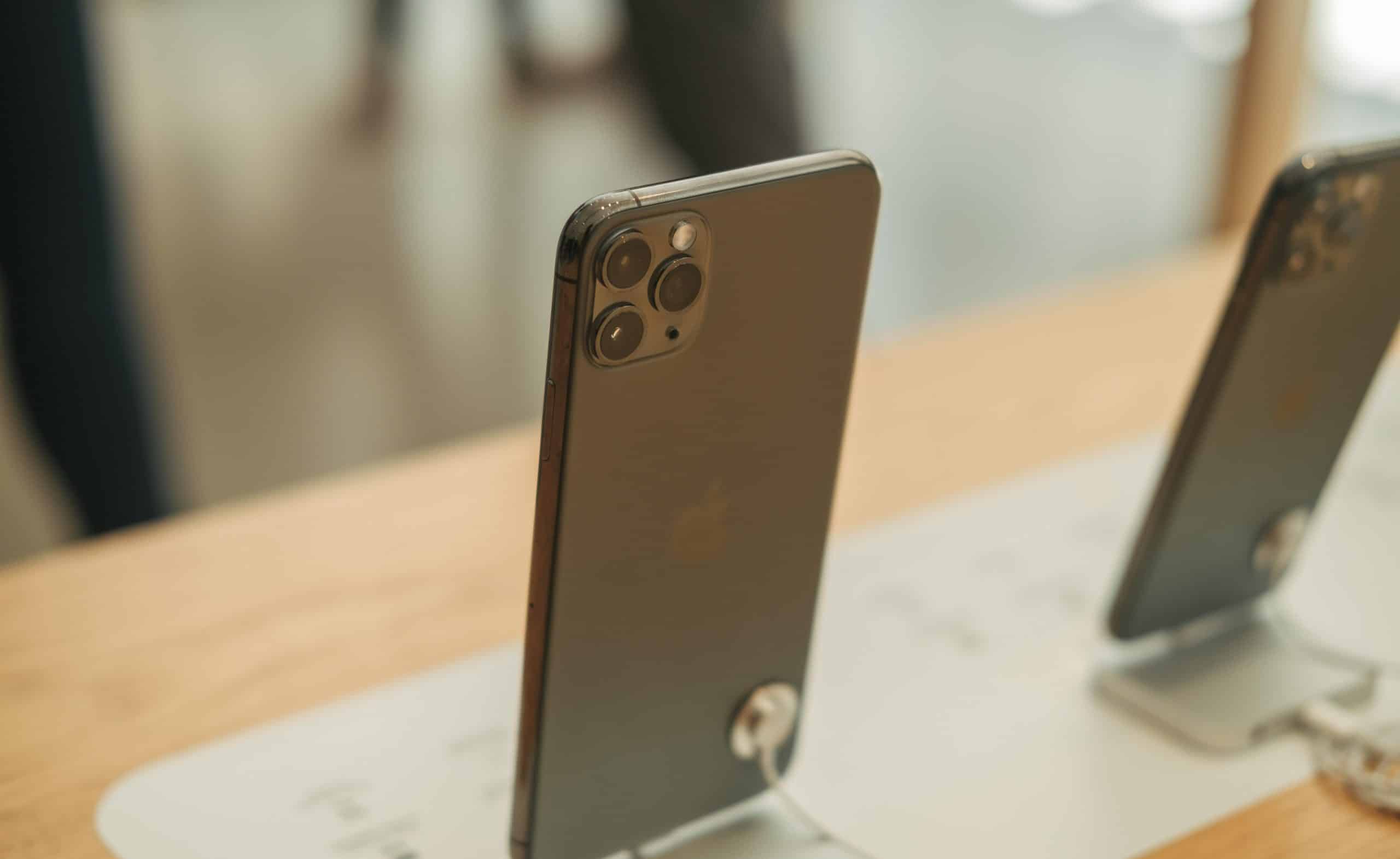 A modern Apple smartphone with a triple-camera setup is displayed on a wooden counter. The phone is standing upright, supported by a security stand. The background is slightly blurred, indicating an indoor environment, likely US stores.