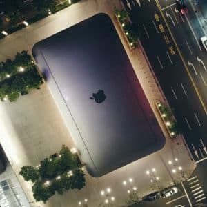 Aerial nighttime view of a modern Apple Store with an Apple logo on its roof, surrounded by well-lit streets and trees. The structure appears sleek and minimalist, embodying the company's signature design aesthetic. Traffic and street markings are visible around the building, promoting their trade-in program.