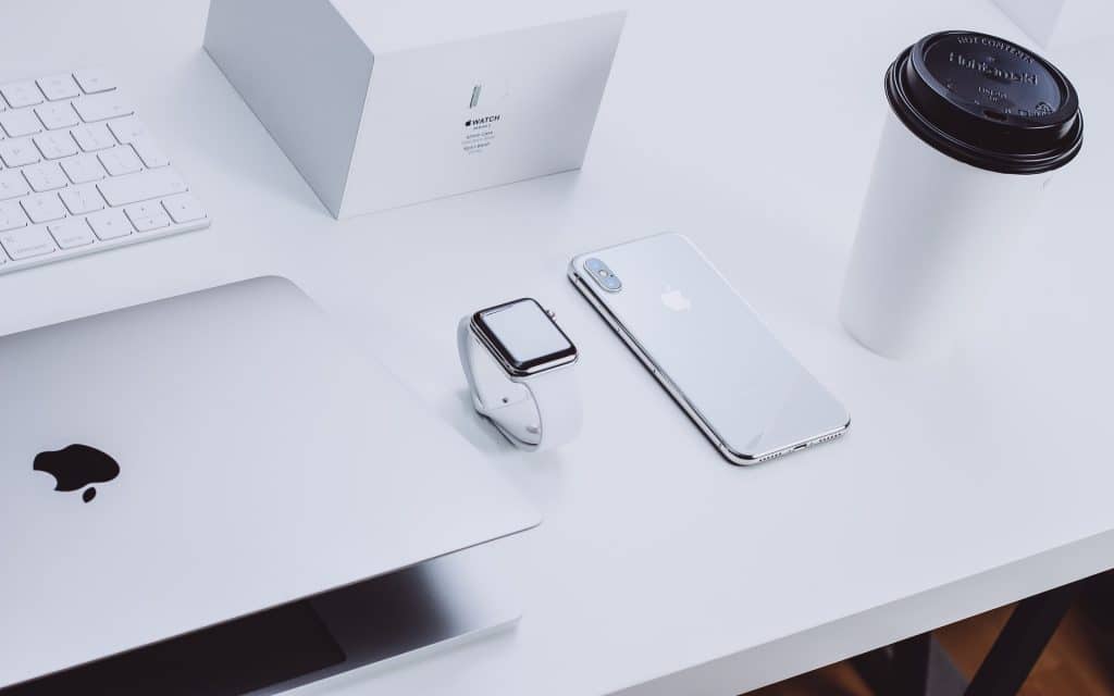 A minimalist workspace featuring a partially open MacBook, an Apple Watch, an iPhone, and a white keyboard, highlighting the elegance of Apple tech. An Apple Watch box and a takeout coffee cup are also neatly arranged on the white desk.