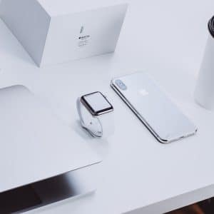 A minimalist workspace featuring a partially open MacBook, an Apple Watch, an iPhone, and a white keyboard, highlighting the elegance of Apple tech. An Apple Watch box and a takeout coffee cup are also neatly arranged on the white desk.