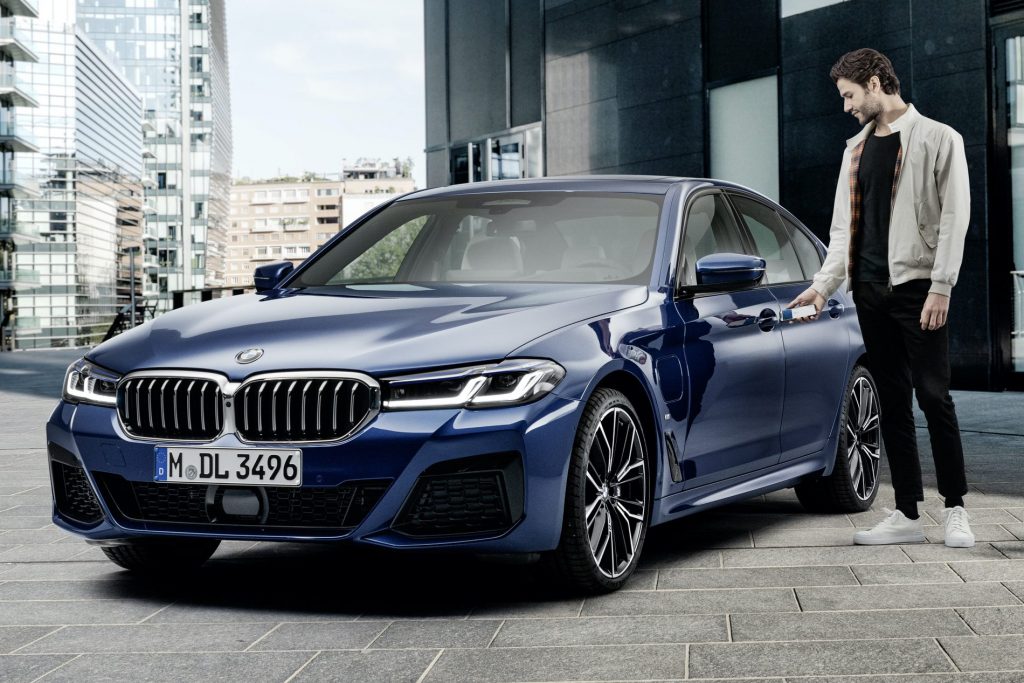 A man dressed in a light jacket, dark pants, and white sneakers stands next to a parked blue BMW sedan in an urban setting, smiling as he reaches for the door handle with his iPhone. The background features modern buildings and a clear sky, highlighting the Apple CarKey compatibility.