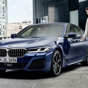 A man dressed in a light jacket, dark pants, and white sneakers stands next to a parked blue BMW sedan in an urban setting, smiling as he reaches for the door handle with his iPhone. The background features modern buildings and a clear sky, highlighting the Apple CarKey compatibility.