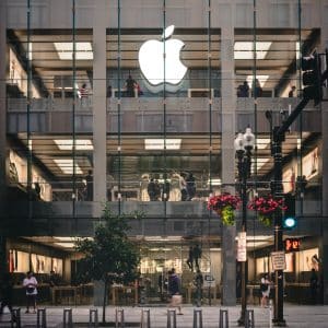 A multi-story Apple Store in the US features a large illuminated Apple logo on the facade. The building has a glass exterior, offering a clear view of the interior bustling with people, despite recent COVID-19 outbreaks. Flower-hanging streetlights and bike racks adorn the sidewalk in front of the store.