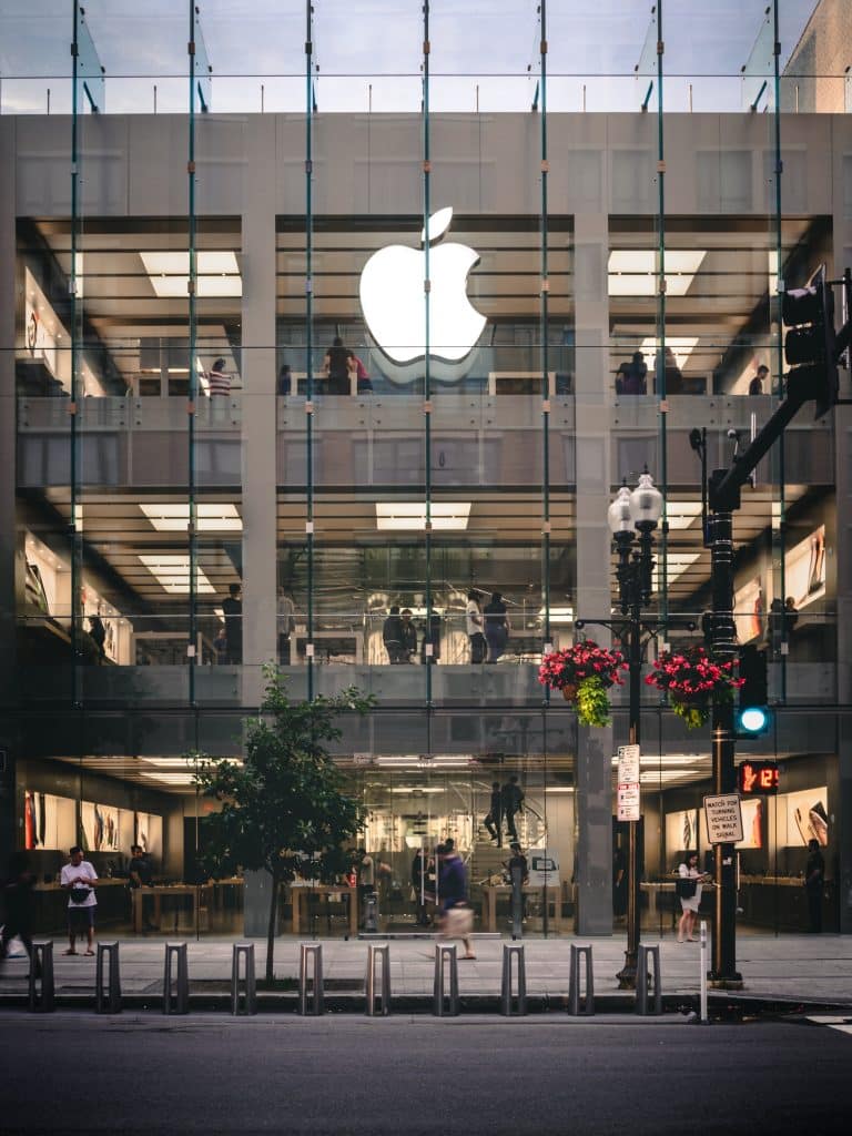 A multi-story Apple Store in the US features a large illuminated Apple logo on the facade. The building has a glass exterior, offering a clear view of the interior bustling with people, despite recent COVID-19 outbreaks. Flower-hanging streetlights and bike racks adorn the sidewalk in front of the store.