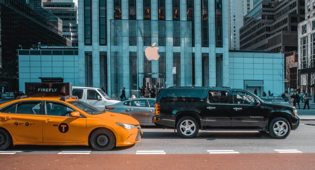 A bustling city street with a yellow taxi and a black SUV in the foreground. In the background, people are walking in front of a tall building adorned with a large Apple logo, signaling an Apple Store entrance—part of the reopening US retail stores wave. Skyscrapers surround the scene.