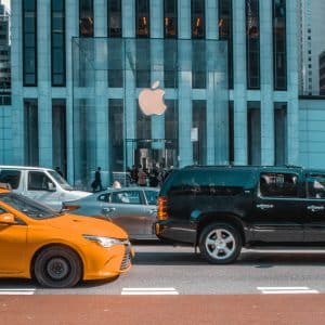 A bustling city street with a yellow taxi and a black SUV in the foreground. In the background, people are walking in front of a tall building adorned with a large Apple logo, signaling an Apple Store entrance—part of the reopening US retail stores wave. Skyscrapers surround the scene.