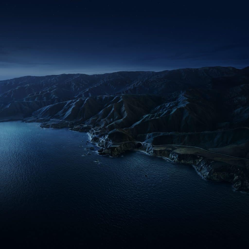 A serene nighttime view of a rugged coastal landscape with dark cliffs meeting the calm, reflective waters of an ocean. The coastline winds along mountainous terrain under a clear, starry sky, creating a tranquil and picturesque scene, reminiscent of the iconic macOS Big Sur wallpapers.