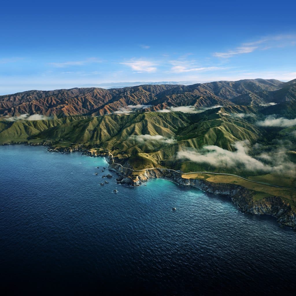 Aerial view of a coastline with lush green hills and mountains under a clear blue sky. Mist drifts over the landscape, and a winding road follows the rugged shoreline. The deep blue ocean contrasts with the vibrant greenery of the land, reminiscent of macOS Big Sur wallpapers designed for your iPhone or Mac.