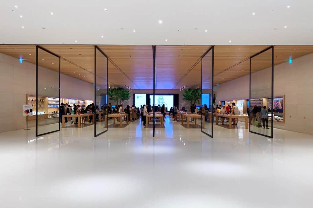 Spacious, modern store with translucent glass doors, wooden ceiling, and neatly arranged tables showcasing electronic products. People are browsing and interacting with the displays. The interior features potted plants and wall-mounted screens, reminiscent of Apple stores in Northern Ireland.