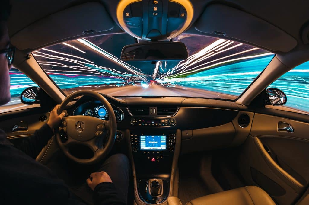 A person is driving a Mercedes-Benz car at night with a view of a streaked, lighted highway ahead. The dashboard and steering wheel are visible, showcasing the car's interior design. With Apple's CarKey feature in iOS 13.6 beta, the light trails create a sense of high speed and motion through the windshield.