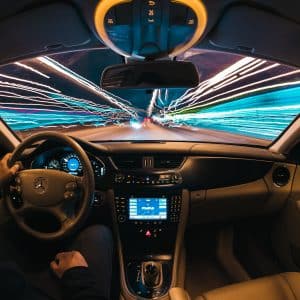 A person is driving a Mercedes-Benz car at night with a view of a streaked, lighted highway ahead. The dashboard and steering wheel are visible, showcasing the car's interior design. With Apple's CarKey feature in iOS 13.6 beta, the light trails create a sense of high speed and motion through the windshield.