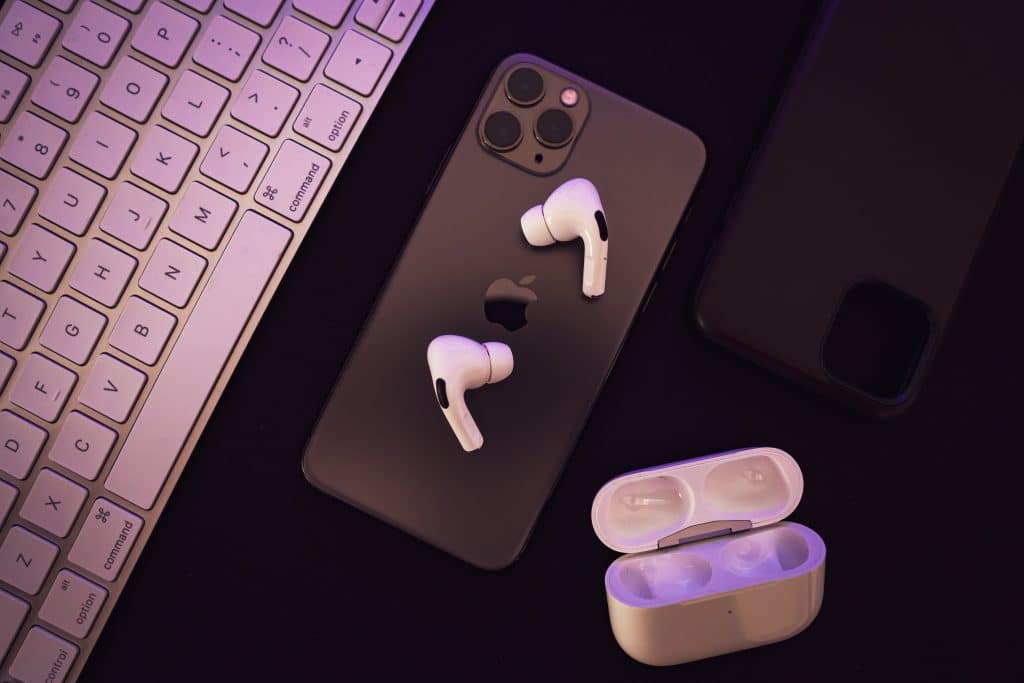 An overhead view of a smartphone, wireless earphones, and a keyboard. The smartphone has two earphones lying on it, while a black phone case is visible nearby. Below is an open AirPods charging case for the earphones.