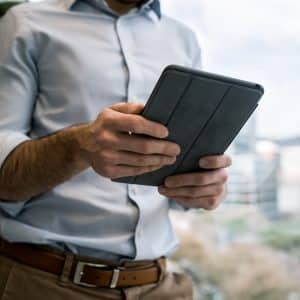 A person wearing a light blue shirt and brown belt is holding and using a tablet. The background features blurred indoor plants and a window showing an out-of-focus cityscape, highlighting the growing trend in tablet sales reported by Canalys.