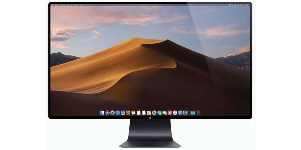 A widescreen computer monitor displays a desktop screen with a desert sand dune landscape under a blue sky. The screen shows various icons and widgets along the bottom. The sleek, black stand hints at the new iMac design unveiled at the recent Apple launch during WWDC.