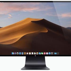 A widescreen computer monitor displays a desktop screen with a desert sand dune landscape under a blue sky. The screen shows various icons and widgets along the bottom. The sleek, black stand hints at the new iMac design unveiled at the recent Apple launch during WWDC.