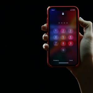 A hand holds a smartphone with a red case against a black background. The phone's screen shows a lock screen with a numeric keypad for entering a passcode, highlighting the importance of password management. The numbers 1 to 9 and 0 are visible, along with options for Emergency and Cancel at the bottom.